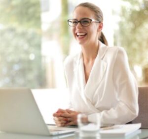 Home 15 Cheerful businesswoman in glasses working on a laptop, in a bright and modern office setting.